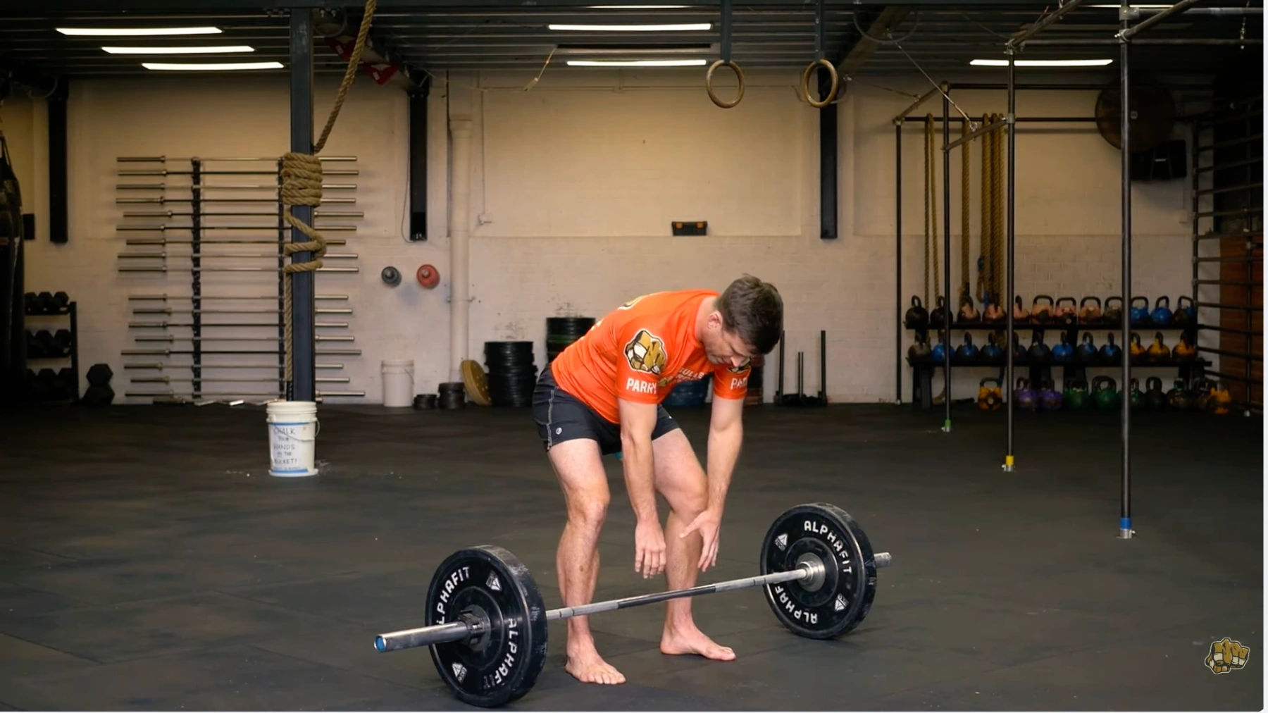 a person in a gym bends down to lift a barbell, barefoot, with kettlebells and ropes visible in the background