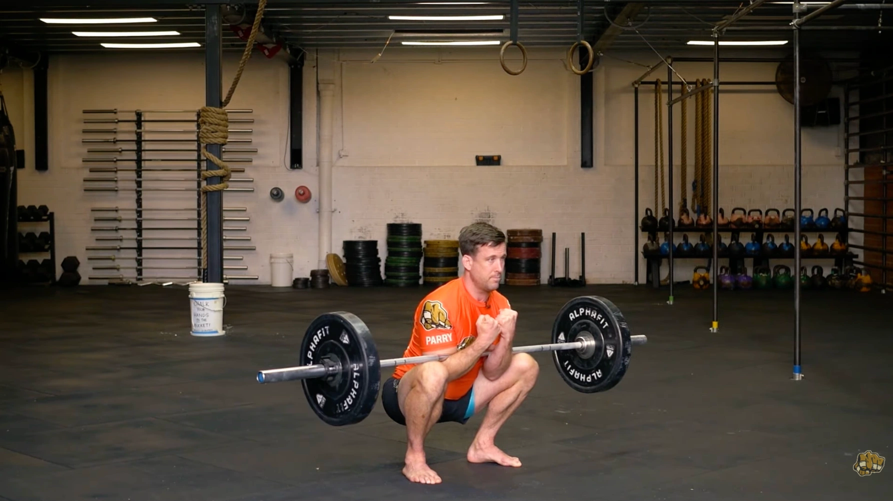 a person performs a zercher squat, holding a barbell at the elbows, in a gym setting with kettlebells in the background