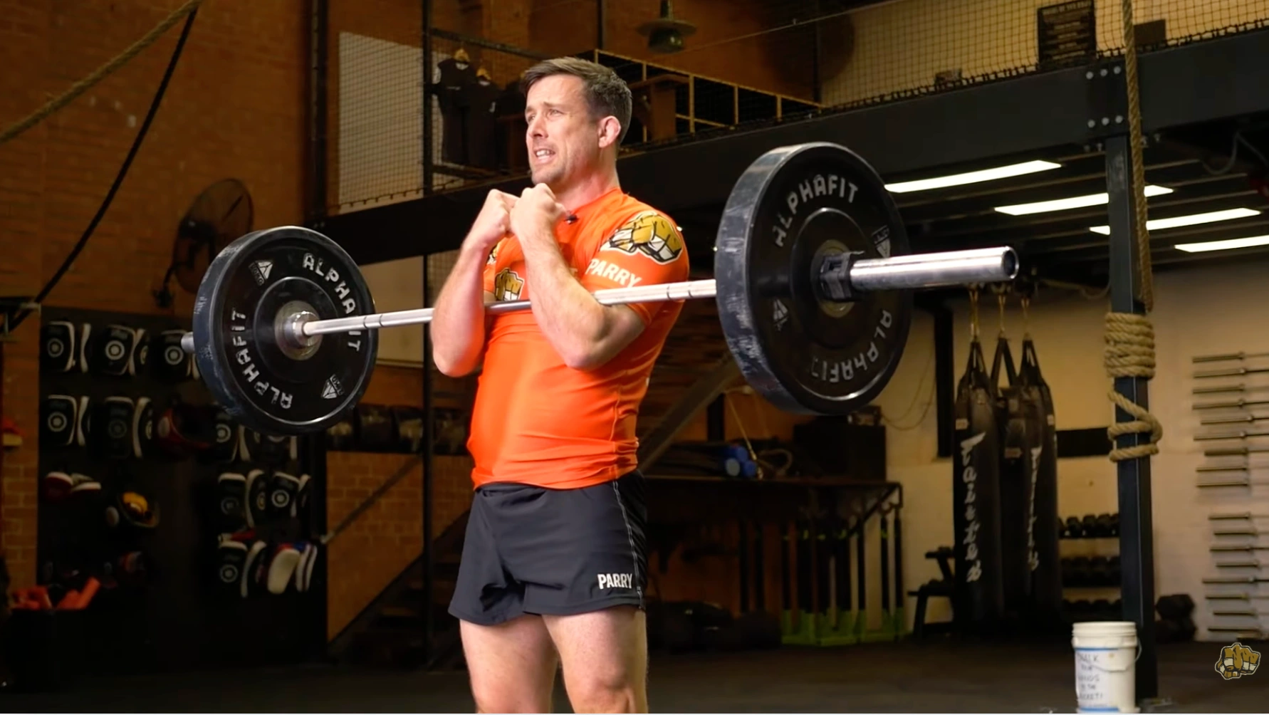a person performs a zercher squat with a barbell racked at the elbows, lifting in a gym with boxing bags