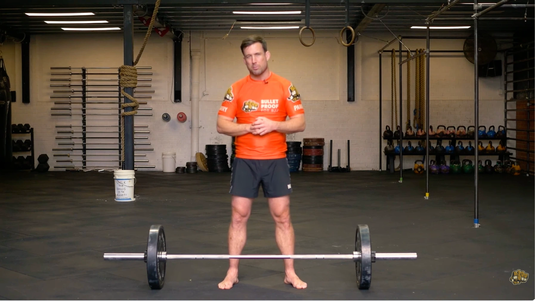 a person stands in a gym next to a barbell with a rack, kettlebells, and rope rings overhead