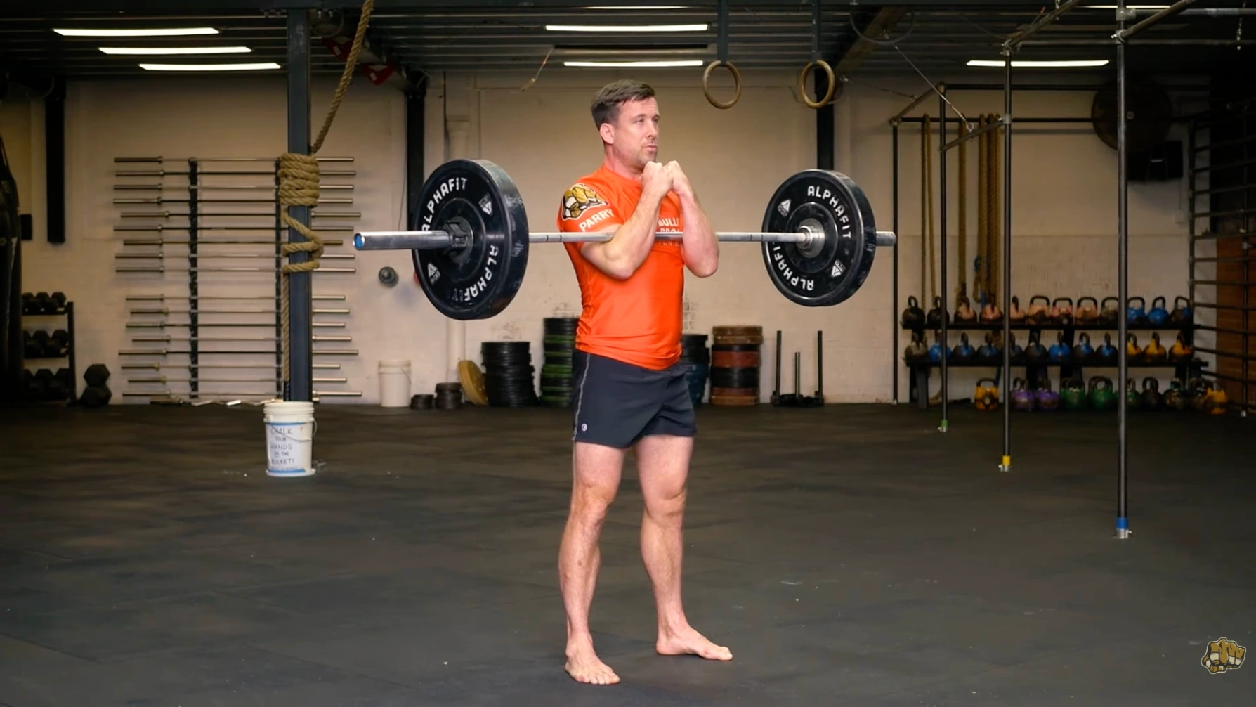a person stands with a barbell racked on their elbows in the front squat position, barefoot, in a gym setting